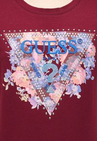 Μπορντώ ύφασμα με σχέδιο λουλουδιών σε τρίγωνο, ασημένιες καρφίτσες και μπλε κείμενο "GUESS U.S.A." που περιβάλλει ένα θαυμαστικό καλυμμένο με στρας.