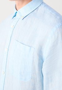 Camicetta button-up in lino azzurro chiaro con colletto, tasca sul petto e tessuto testurizzato. Presenta bottoni bianchi lungo la parte anteriore.
