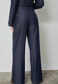 Sötétkék, széles szárú farmernadrág. Hátul kontrasztos varrással ellátott zsebekkel rendelkezik, és sima, strukturált sziluettet mutat.