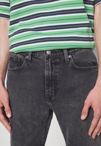 Jeans in denim nero con finitura testurizzata, caratterizzati da una chiusura con bottoni e tasche laterali, indossati con una camicia verde a righe.