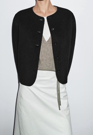 Kobieta w czarnym kardiganie na guziki, beżowym topie kopertowym i białej spódnicy, sfotografowana na jednolitym białym tle.