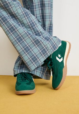 Personne portant des baskets en daim vert avec un logo étoile blanc et un pantalon large à carreaux bleu clair, debout sur un sol jaune.