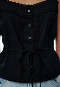 Haut en coton noir avec bordure en dentelle et fermeture à boutons sur le devant. Comprend une ceinture à nouer et des plis verticaux, créant une silhouette ajustée.