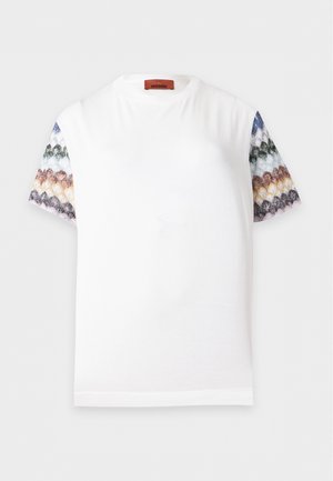 Femme portant des lunettes de soleil noires, un t-shirt blanc avec des manches à motifs multicolores, un pantalon gris évasé et des baskets claires, debout avec une main sur la hanche.