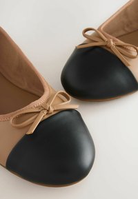Balletflats met een tan leren bovenkant en een ronde zwarte leren neuskap, verfraaid met een kleine bijpassende strik op elke schoen.