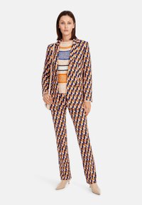 Musterhosen und Blazer in Blau, Orange und Creme, mit einem geometrischen Design. Das Model trägt einen strukturierten Pullover und minimalistische Schuhe.