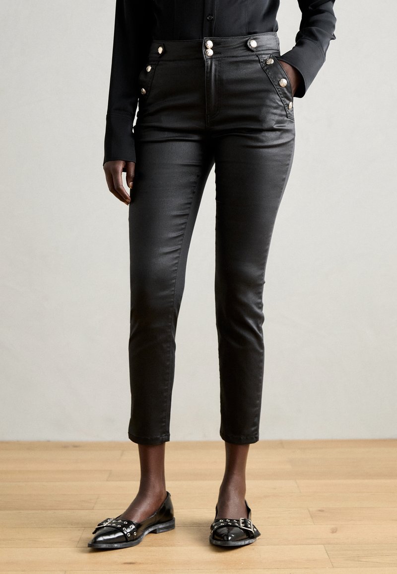 Personne debout sur un plancher en bois, portant un pantalon noir coupé court avec des boutons décoratifs, une chemise noire et des mocassins noirs ornés.