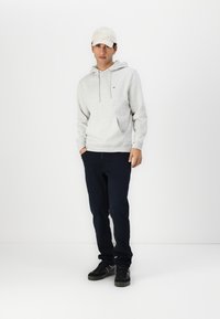 Grijze hoodie met een voorzak en capuchon, gecombineerd met donkerblauwe broek en zwarte sneakers. De hoodie heeft een klein logo op de borst.