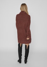Abito maglione di color ruggine con colletto alto e spacchi laterali, abbinato a stivali neri al ginocchio con design a taglio.