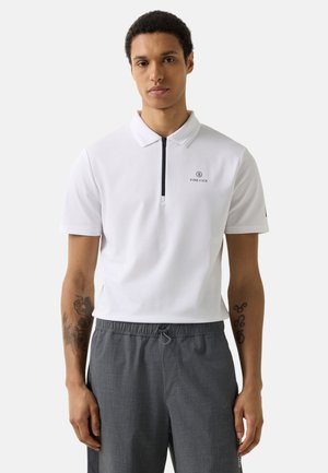 Mann trägt weißes kurzärmeliges Polo mit schwarzem Reißverschluss und grauen Kordelzughosen, steht vor einem schlichten hellen Hintergrund.