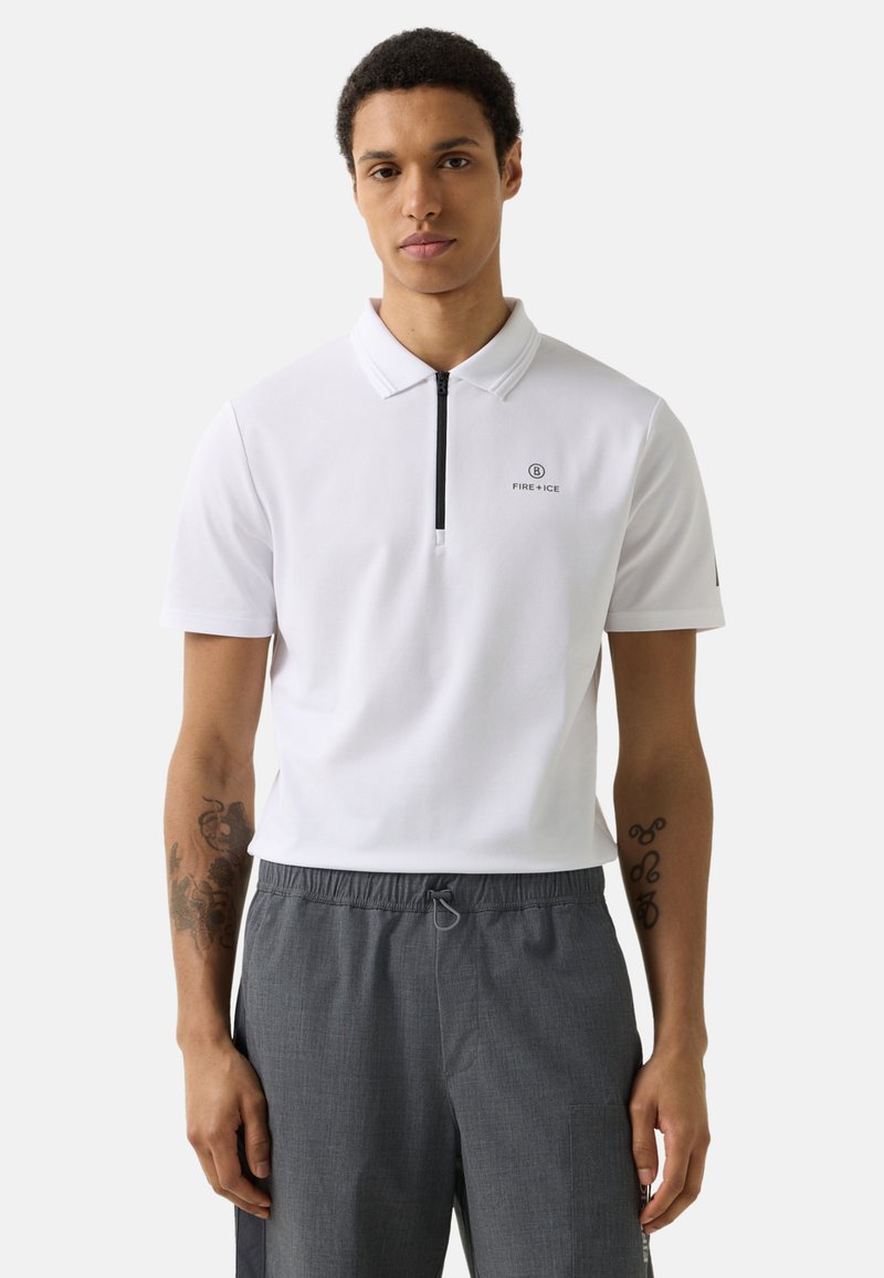 Man wearing white short-sleeve polo with black zipper and gray drawstring pants, standing against plain light background.