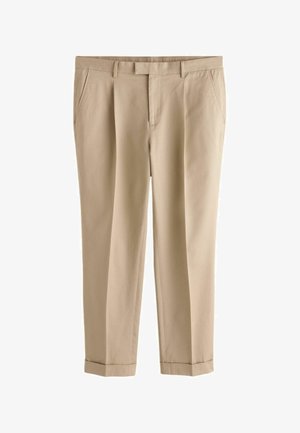 Bărbat purtând o cămașă albastru deschis, cravată bleumarin, pantaloni bej și pantofi negri, stând în picioare lângă un perete bej și privindu-se spre dreapta.