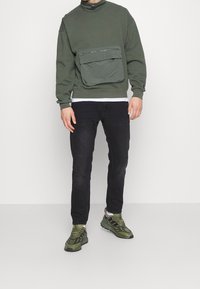 Sudadera verde oliva con cuello alto y un gran bolsillo delantero, combinada con jeans negros y zapatos deportivos verdes, con un acabado texturizado.