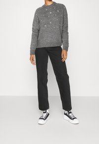 Grau gestrickter Pullover mit Perlenakzenten, ribbelnden Bündchen, kombiniert mit schwarzen, geraden Jeans und schwarz-weißen Leinen-Sneakern.