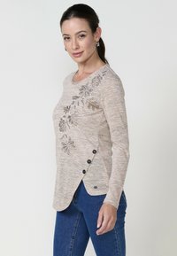 Beige Langarmshirt mit floralem Stickerei am Schulter, Seitenschaltern und einem abgerundeten Saum. Strukturiertes Material mit lässiger Passform.
