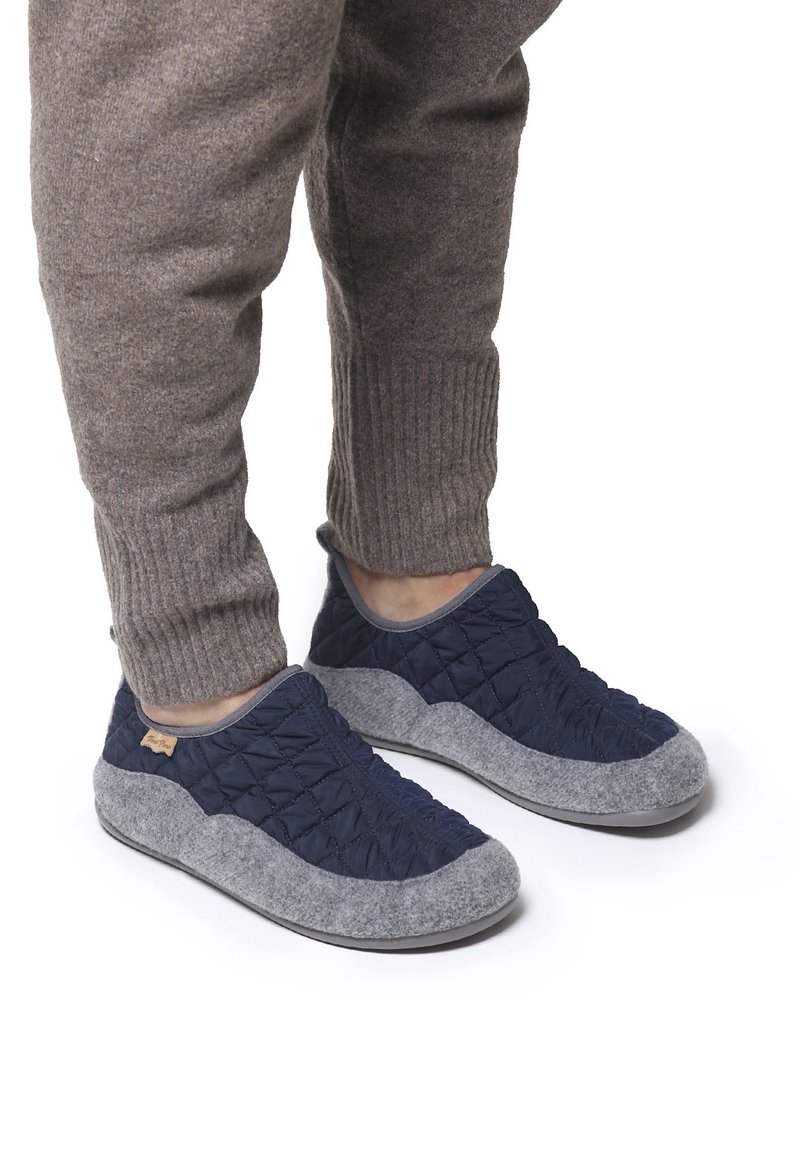 Personne portant des chaussons matelassés bleu marine et gris, associés à un pantalon de jogging marron en maille côtelée, sur fond blanc.