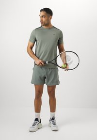 T-shirt atlético verde com mangas curtas, calções a condizer e ténis brancos. Segurando uma raquete de ténis com uma bola. Design minimalista.