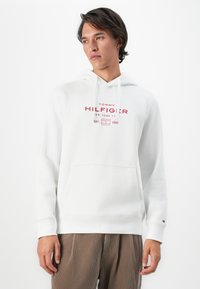 Jeune homme portant un sweat à capuche blanc Tommy Hilfiger avec un logo rouge et un pantalon en velours côtelé marron, se tenant contre un fond blanc uni.