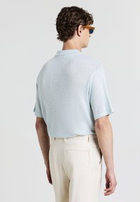 Mann, der ein hellblaues kurzärmliges Strickhemd und beige Hosen trägt, von hinten vor einem einfarbigen hellgrauen Hintergrund gesehen.