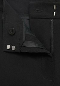 Pantalon noir avec une braguette zippée, une fermeture à bouton et des accents métalliques. Le tissu semble lisse avec un léger aspect brillant.