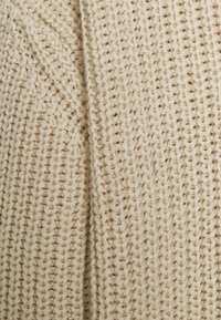 Beige stickat tyg med ett texturerat mönster och synliga stygn. Vävnaden är lös, vilket framhäver de naturliga fibrerna som används.