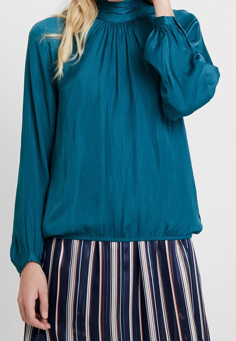 Blouse longue à manches teal avec un col montant, des détails froncés et une texture douce ; les manches sont légèrement bouffantes et les poignets sont boutonnés.