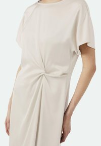 Beżowa sukienka z gładkiego materiału, z okrągłym dekoltem, krótkimi rękawami i detalem w formie skrętu na talii, podkreślona miękkimi fałdami.