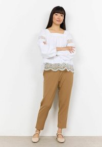 Hvid bluse med blondekant, afslappet pasform og pufærmer, parret med lysebrune tætsiddende bukser. Beige flade sko fuldender outfittet.