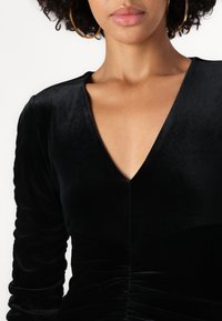 Zwarte fluwelen jurk met een V-hals, lange mouwen en geplooide details bij de taille. Zachte textuur met een aansluitende silhouet.