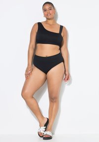 Set bikini nero a coste con top a spalla unica e parte inferiore a vita alta. La modella indossa sandali slide bianchi e neri. Texture liscia.