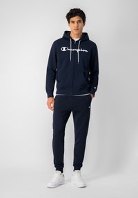 Sudadera con cremallera de color navy con el logotipo blanco de "Champion", combinada con pantalones a juego, ambos de tela suave. Incluye bolsillos frontales y capucha con cordón.