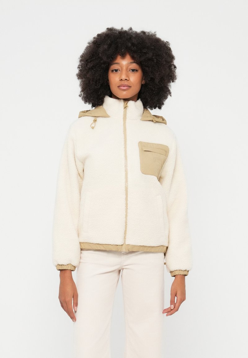 Chaqueta de forro polar crema con detalles beige y un bolsillo delantero. Cuenta con un cuello alto, puños elásticos y cierre de cremallera.