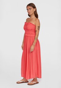 Abito maxi senza spalline color corallo con bustier arricciato e dettagli volant, in tessuto fluido, indossato con infradito.