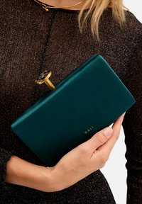 Teal sammet clutchväska med rektangulär form, guldhardware och ett runt handtag med ett svart och vitt mönster. "NALI" präglat i guld.