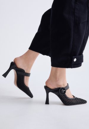 Zapatos mule de malla negra con tacón alto, punta afilada y tiras con hebilla, usados con pantalones negros recortados sobre un fondo claro.