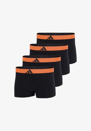 Umięśniony mężczyzna stojący boso, ubrany w czarne bokserki z pomarańczowym pasem w talii ozdobionym czarnym logo Adidas.