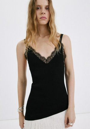 Jeune femme aux cheveux blonds ondulés portant un haut noir sans manches orné de dentelle et une jupe blanche texturée, debout devant un fond uni.