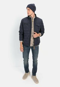 Marineblå quiltet jakke med stående krave, lynlås fortil og brystlommer, båret over en beige sweater og matchet med jeans og sneakers.