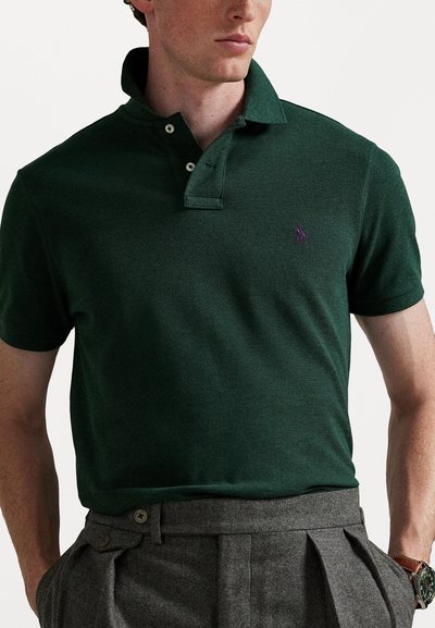 Cămașă polo verde închis, cu un finisaj texturat, guler cu două butoane și accent de logo violet. Asortată cu pantaloni gri plisati.