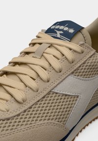 Μπεζ αθλητικό παπούτσι με πάνελ από δίχτυ και σουέτ, μπεζ κορδόνια και σκούρο μπλε ψηφιακό ετικετάκι στη γλώσσα με λευκή στυλιζαρισμένη επιγραφή της μάρκας "Diadora".