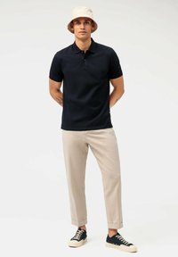 Marineblaue Poloshirt mit kurzen Ärmeln und Kragen, kombiniert mit hellbeigen Hosen und schwarzen Sneakers mit cremefarbenen Sohlen. Bucket Hat inklusive.