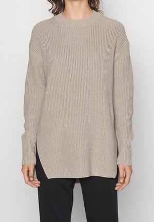 Beżowy, prążkowany sweter z dzianiny z długimi rękawami i okrągłym dekoltem, noszony przez osobę o kręconych włosach, zestawiony z czarnymi spodniami.