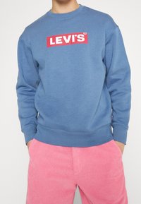 Persona che indossa una felpa blu con logo rosso e bianco della Levi's e pantaloni di velluto a coste rosa, con le mani nelle tasche.