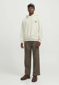Felpa crema con cappuccio e coulisse, abbinata a pantaloni brown larghi e scarpe brown con lacci; berretto blu in testa.