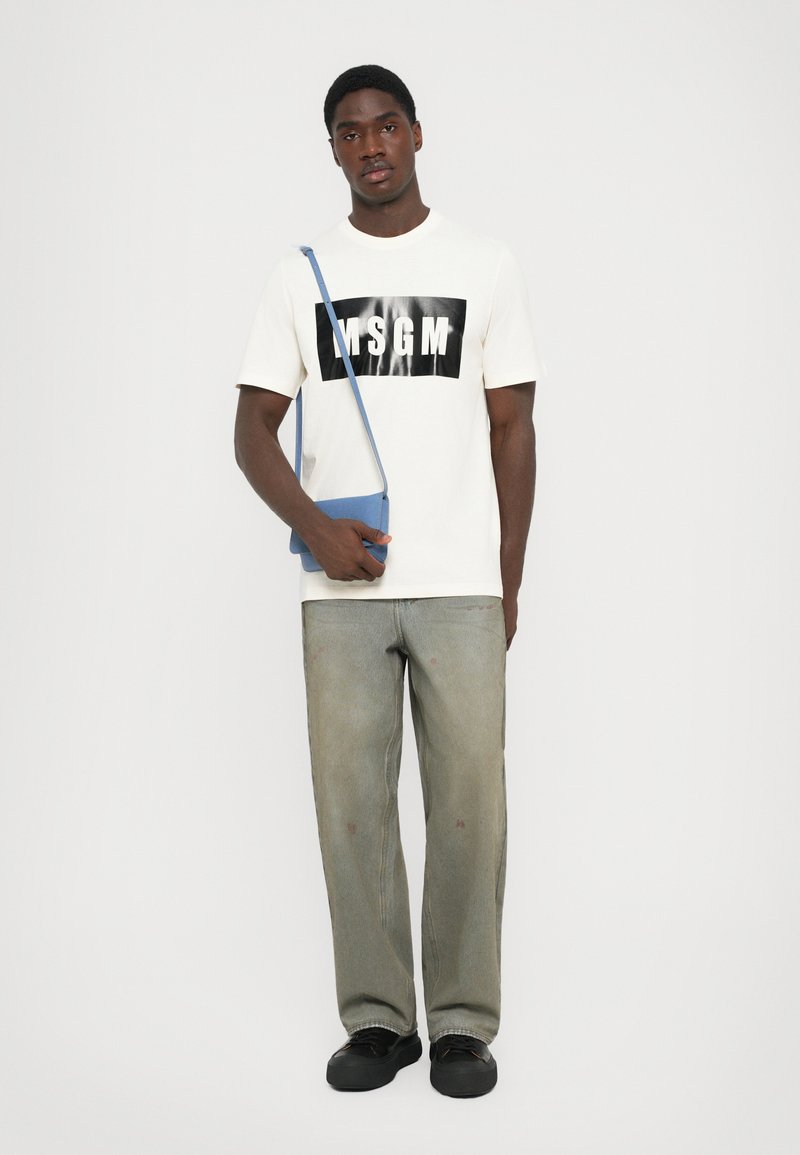 White cotton t-shirt with black "MSGM" logo, paired with loose light green jeans and black sneakers. Blue crossbody bag adds color contrast.
