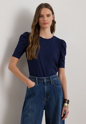 Temno modra majica s kratkimi rokavi in nabranimi rameni, kombinirana z visokim pasom modrih jeans hlač. Zapestnica kot dodatek na zapestju.