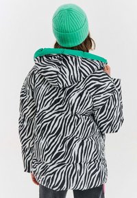 Osoba oblečená v čierno-bielej pufovacej bunde so zebrovým pruhom, s zeleným podšívkou kapucne a jasne zelenou pletenou čiapkou, stojí chrbtom.