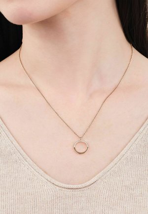 Delicado collar de cadena de oro con un colgante circular que presenta pequeños cristales incrustados, llevado en el cuello de una mujer sobre un top de punto beige.
