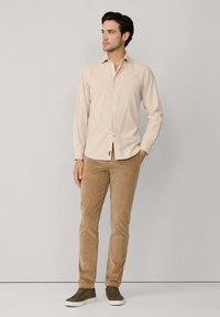 Chemise beige clair à boutons, manches longues, en tissu lisse. Associée à un pantalon en velours côtelé beige et des baskets olive, se tenant contre un fond gris.