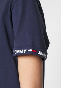 Mörkblå bomullströja med en standard passform. Utrullade ärmar visar "TOMMY" och "JEANS" i vit text tillsammans med en röd och vit accentlinje.